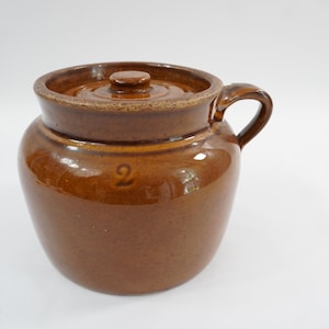 May include: A brown glazed ceramic crock with a matching lid and a single side handle. The body of the crock features a stamped number 2 on the front. The piece has a smooth, glossy finish and a rounded shape with a wide opening and a flat lid.