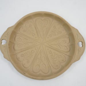 May include: A round, light tan stoneware baking mold featuring a radial pattern of eight heart-shaped sections, each embossed with a floral design. The mold includes two side handles with small heart-shaped cutouts for easy gripping and handling.