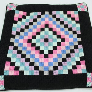May include: Square black cushion with a woven checkerboard pattern in pink, blue, grey, and black. The corners have a small floral design in pink and blue. The cushion is likely for home decor.