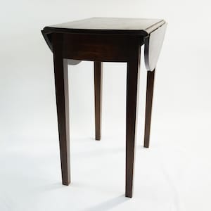 May include: A dark brown, small drop-leaf table with a rectangular top and four slender legs. The table's leaves are folded down. The table is approximately 61 cm tall.