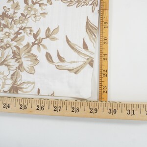 May include: Close-up of a white fabric with a floral pattern in shades of beige and brown. A wooden ruler is placed along the edge of the fabric, showing measurements in inches. The fabric has a vintage or antique aesthetic.