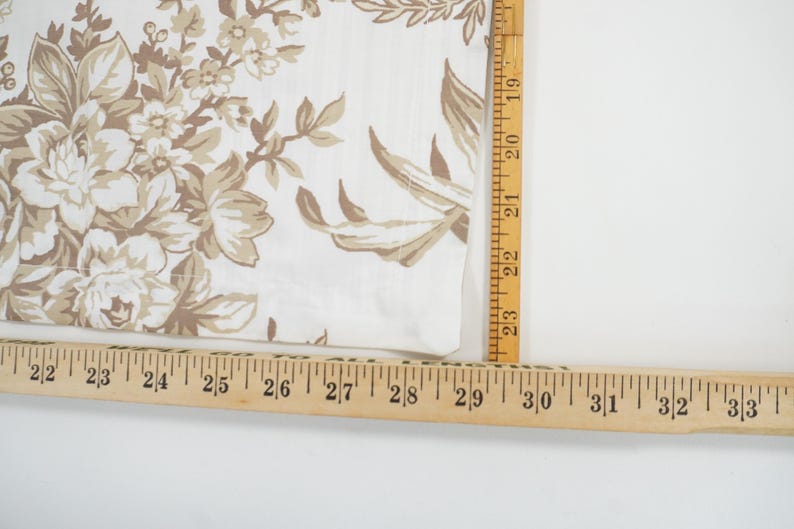 May include: Close-up of a white fabric with a floral pattern in shades of beige and brown. A wooden ruler is placed along the edge of the fabric, showing measurements in inches. The fabric has a vintage or antique aesthetic.