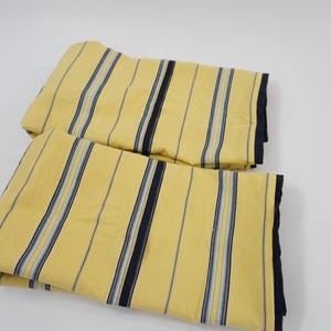 Può includere: Due pannelli di tessuto piegati caratterizzati da un colore di base giallo con strisce verticali nelle tonalità blu navy, azzurro e bianco. Il tessuto ha una consistenza liscia ed è disposto in due pile rettangolari piegate ordinatamente.