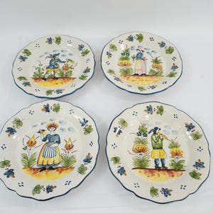 May include: A set of four round ceramic plates with scalloped edges, each featuring a hand-painted illustration of a figure in traditional clothing surrounded by stylised flowers and foliage in shades of blue, yellow, green, and brown on a cream base.