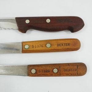May include: Three kitchen knives with wooden handles and stainless steel blades. The top knife has a serrated edge. The handles are brown and tan, with the word "DEXTER" printed on them.