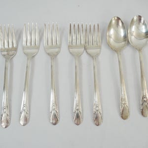 May include: A set of seven silver-toned metal utensils consisting of five forks and two spoons. Each piece features a slender handle with a decorative embossed floral pattern at the base and a polished finish, arranged in a row on a plain surface.