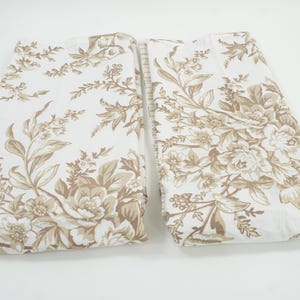 May include: Two folded fabric pieces with a floral pattern. The design features large, detailed flowers and foliage in shades of brown and tan against a white background. The fabric appears to be cotton or a similar material.
