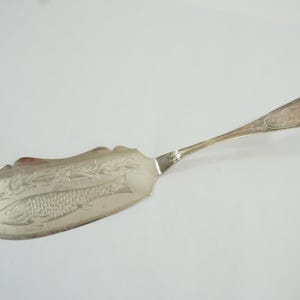 May include: Silver fish serving utensil with a decorative handle and a wide, curved blade. The blade features an etched fish design and floral accents, perfect for serving seafood dishes.