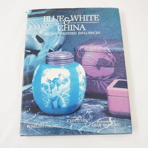 May include: A book titled "Blue & White China: Origins/Western Influences" with a blue and white ceramic jar on the cover. The jar has a lid and a detailed design. Other objects in the background are a purple box and a pink box.