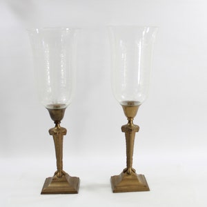 Puede incluir: Un par de candelabros de metal dorado con pantallas de huracán de vidrio transparente. Los candelabros tienen una base cuadrada y un diseño decorativo.