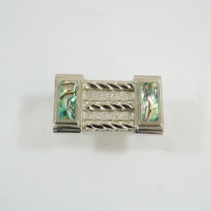 May include: Silver-toned metal barrette with a rectangular design. The barrette features a central section with a twisted rope pattern and two end pieces with iridescent abalone shell inlays. A decorative hair accessory.