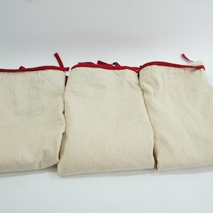 Peut inclure: Trois sacs de rangement en tissu de couleur crème avec une bordure rouge. Les sacs sont pliés et empilés les uns sur les autres.