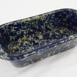 May include: A rectangular loaf pan featuring a deep blue and cream-colored speckled sponge-painted pattern. The ceramic bakeware has rounded corners, a wide rim, and a small white rectangular sticker with the number 6 on the left handle.
