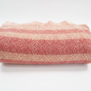 May include: Folded woven textile in shades of red and beige. The fabric features horizontal stripes of varying widths and textures. The textile is likely a blanket or throw, with a soft, inviting appearance. The color palette is warm and neutral.