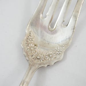 May include: A silver-toned metal fork featuring a decorative embossed pattern of small daisy-like flowers at the base of the tines. The handle transitions into a wide, scalloped neck area where the floral design is centered on the front surface.