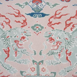 May include: A needlepoint textile featuring a light pink background with two symmetrical light green mythical lion figures facing each other. The design includes red flame-like accents, stylized blue and green clouds, and geometric floral motifs.