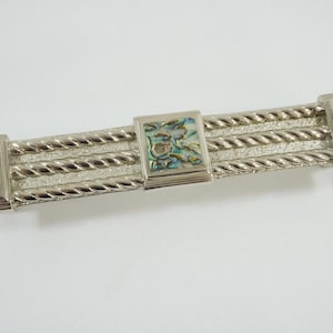 Könnte beinhalten: Silberfarbene Haarklammer mit geflochtenem Design. Die Haarklammer hat rechteckige Abalone-Muschel-Inlays an jedem Ende und in der Mitte. Die Abalone-Muschel hat schillernde blaue, grüne und braune Farbtöne.