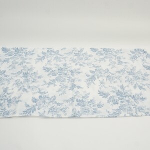 May include: A rectangular, white fabric with a repeating blue floral pattern. The design features detailed flowers and foliage, creating a classic, elegant aesthetic. The fabric is laid flat against a white background.
