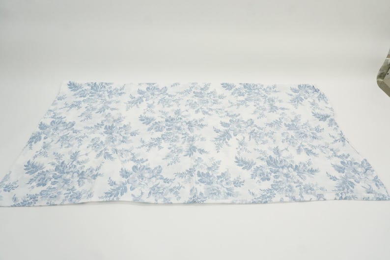May include: A rectangular, white fabric with a repeating blue floral pattern. The design features detailed flowers and foliage, creating a classic, elegant aesthetic. The fabric is laid flat against a white background.
