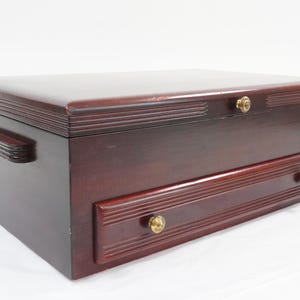Puede incluir: Un cofre de almacenamiento rectangular de madera color caoba oscuro con una tapa abatible con detalles de ranuras horizontales, un cajón frontal con dos pequeños pomos redondos de tono latón y asas laterales ranuradas a juego.