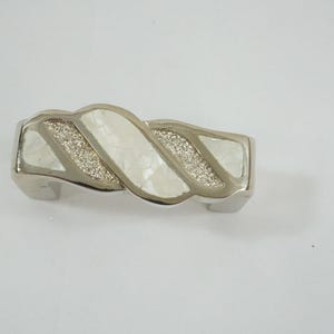 Könnte beinhalten: Silberfarbenes Metallarmband mit einem gedrehten Design. Das Armband hat Abschnitte aus weißem, schillerndem Material und Abschnitte aus glitzerndem Material. Das Armband wird auf weißem Hintergrund gezeigt.