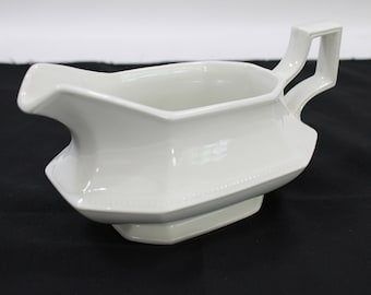 Może przedstawiać: Biały ceramiczny sosjerka o gładkim, błyszczącym wykończeniu, z szerokim dzióbkiem i geometrycznym, kanciastym uchwytem. Podstawa jest prostokątna ze ściętymi krawędziami, a korpus posiada subtelną, wypukłą fakturę koralików wzdłuż dolnej krawędzi.