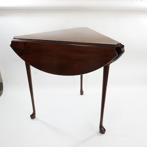 May include: A dark brown, antique-style, three-legged table with a triangular top and a rounded, drop-leaf side. The table's legs have a curved design. The table is made of wood and has a glossy finish.