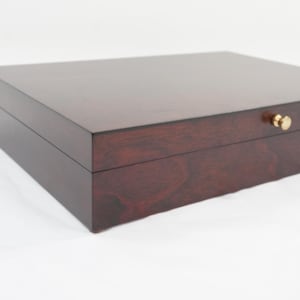 May include: A rectangular wooden storage box with a dark reddish-brown finish and visible wood grain. The box features a smooth, flat lid and a small, round, metallic gold-colored knob centered on the front edge for opening the lid.