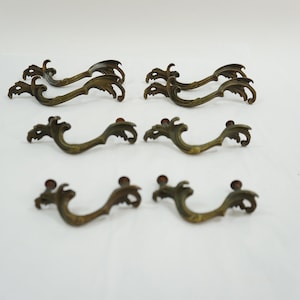 May include: Set of eight antique brass drawer pulls with a decorative, ornate design. Each pull features a stylised, swirling pattern reminiscent of a dragon or a mythical creature.