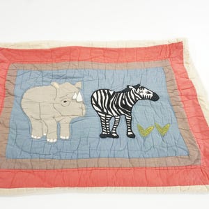 May include: A square, quilted blanket featuring a light blue center with a rhino and zebra applique. The border is red and cream, with a brown accent. The blanket is designed for a nursery or child's room.