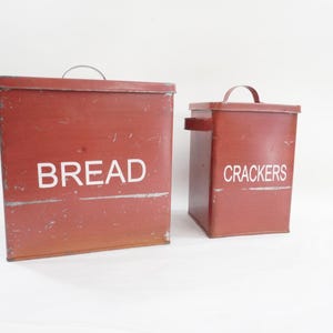 Może przedstawiać: Dwa czerwone metalowe pojemniki do przechowywania chleba i krakersów. Większy pojemnik ma napis "BREAD" białymi literami, a mniejszy napis "CRACKERS". Oba mają uchwyty i są prostokątne.