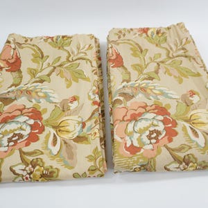 以下が含まれることがあります： 花柄の2つの折り畳まれた布パネル。デザインは、ベージュの背景に、ピーチ、赤、クリームの色合いの大きな様式化された花を特徴としています。緑と茶色の葉が花の要素を強調しています。