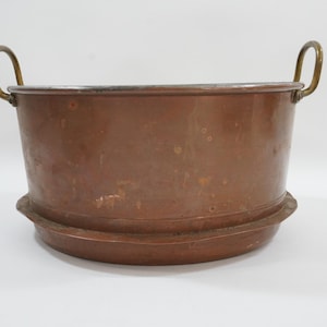 Peut inclure: Une cuve en métal de couleur cuivre avec deux poignées en laiton. La cuve a une forme cylindrique large avec une base légèrement évasée. Les poignées sont arquées et fixées au bord. Probablement vintage.