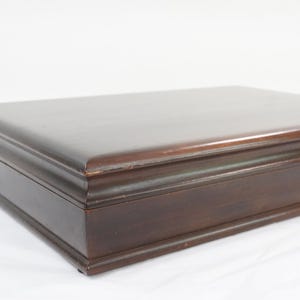 Puede incluir: Una caja rectangular de madera de color marrón oscuro con un acabado liso y pulido. La tapa presenta bordes biselados y una superficie superior plana y limpia. La caja tiene un diseño estructurado y bajo con vetas de madera visibles y sutiles signos de desgaste en las esquinas.
