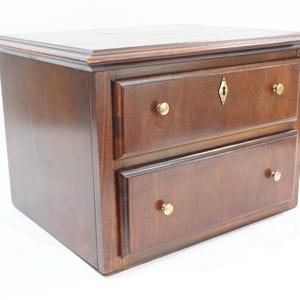 Puede incluir: Un pequeño cofre de madera de color marrón oscuro con dos cajones horizontales. Cada cajón cuenta con dos pomos redondos de latón. El cajón superior incluye un escudo de cerradura central de latón. El cofre tiene una superficie superior plana.