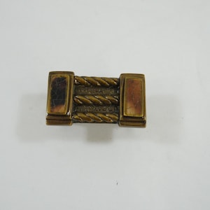May include: A rectangular, antique-style metal clasp with a bronze finish. The clasp features a central section with three twisted rope designs and two end pieces with rectangular insets. The overall design is symmetrical.