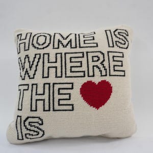 Puede incluir: Cojín decorativo color crema con la frase "HOME IS WHERE THE HEART IS" en letras negras. Un corazón rojo está centrado debajo del texto. El cojín cuadrado tiene una superficie texturizada.