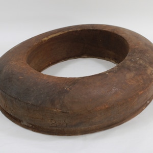 May include: A circular, wooden object with a dark brown finish. The object has a thick, rounded rim and a hollow center. The wood grain is visible. Numbers are stamped on the side. The object appears to be an antique hat form.
