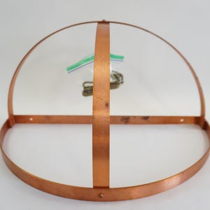 May include: A copper-colored metal frame, possibly for a lampshade or decorative element, with a half-circle top and a horizontal bar. A vertical bar intersects the horizontal bar. A small plastic bag with hardware is visible inside the frame.