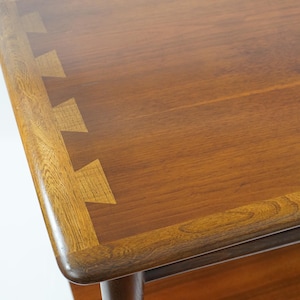 May include: A rectangular wooden table with a smooth, polished brown surface. The edge of the tabletop features a decorative inlay pattern consisting of light-colored, interlocking dovetail-shaped wooden pieces set into the darker wood grain.