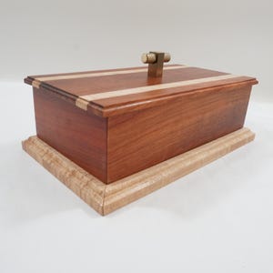 May include: A handcrafted wooden box with a rectangular shape. The box is made of reddish-brown wood with a light-colored wood accent on the lid. It has a brass handle and sits on a light-colored wooden base. The box measures approximately 8 inches long, 4 inches wide, and 3 inches high.