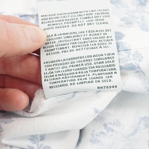 May include: A white fabric tag with care instructions in English, French, and Spanish. The tag is held by a hand and is attached to a white fabric with a blue floral pattern. The tag includes the text "RN76948".