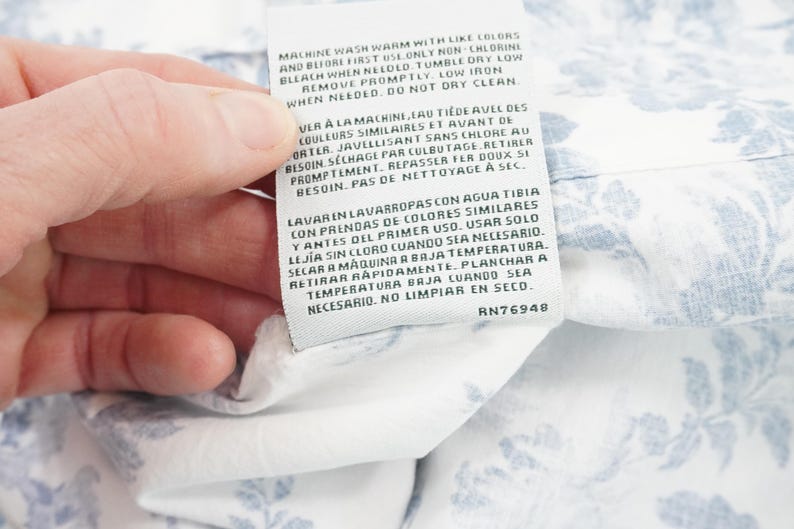 May include: A white fabric tag with care instructions in English, French, and Spanish. The tag is held by a hand and is attached to a white fabric with a blue floral pattern. The tag includes the text "RN76948".