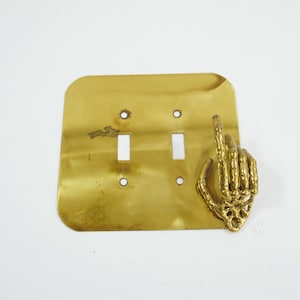 Könnte beinhalten: Eine Lichtschalterabdeckung aus Messing mit zwei Schalteröffnungen und einer dekorativen Hand, die den Mittelfinger zeigt. Die Abdeckung ist rechteckig mit abgerundeten Ecken.