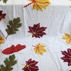 May include: A white textured blanket featuring a repeating pattern of appliqued autumn leaves in shades of deep red, golden yellow, and forest green. The leaves are arranged across the fabric surface, showcasing various shapes including maple and oak.