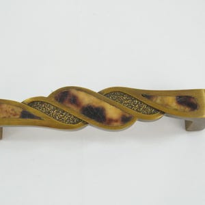 Könnte beinhalten: Dekorative Schrankgriff aus Messing. Der Griff hat ein geflochtenes Design mit Schildpattmuster und einem strukturierten, goldfarbenen Mittelteil. Der Griff ist für eine einfache Installation konzipiert und verleiht Eleganz.