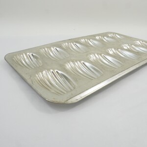 May include: A rectangular silver-colored metal baking pan featuring twelve shell-shaped indentations arranged in two rows. The tray has a smooth, flat border surrounding the molded cavities, which are designed for baking traditional madeleine cakes.