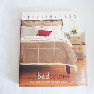 May include: A book titled "bedrooms" by Pottery Barn. The cover features a photograph of a bed with a tan duvet cover and white and tan pillows. The bed is in a bedroom with a wooden floor and a small wooden table with a lamp on it.