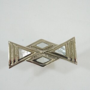 May include: Silver-toned metal brooch with a geometric design. The brooch features a central diamond shape and two triangular shapes on either side, with iridescent inlays. The brooch is set against a white background.