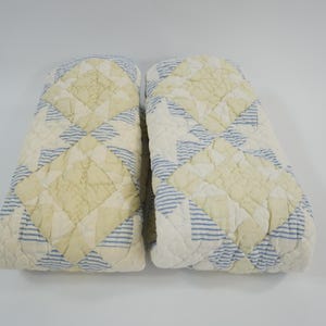 May include: Two folded quilted blankets featuring a geometric pattern of cream-colored squares and blue and white striped triangles. The fabric has a visible stitched texture throughout, creating a repeating diamond and star-like design across both pieces.
